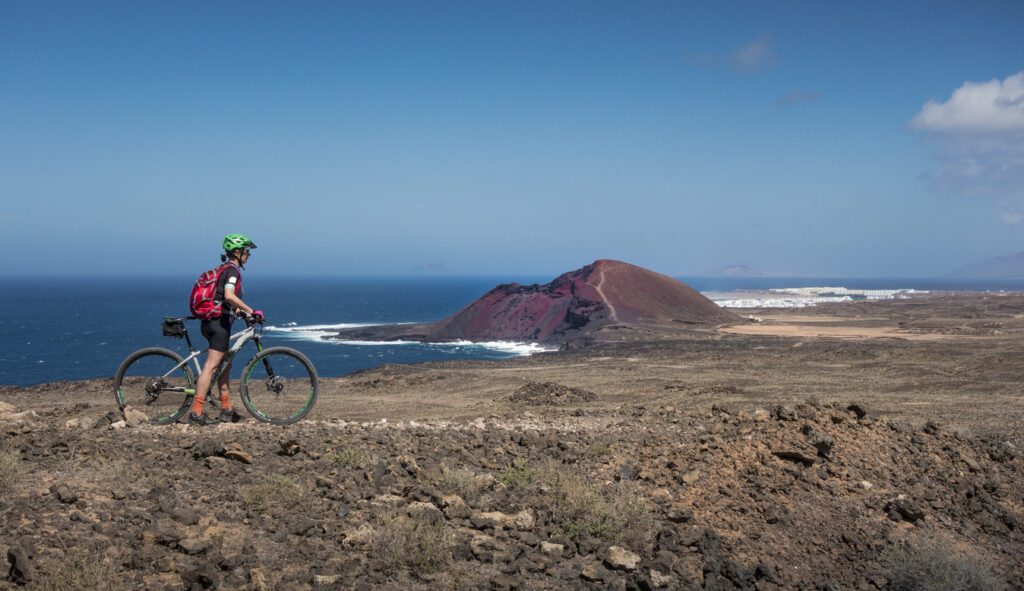 Radfahrer steht mit Mountainbike auf karger Vulkanlandschaft und blickt auf Meer und roten Hügel.