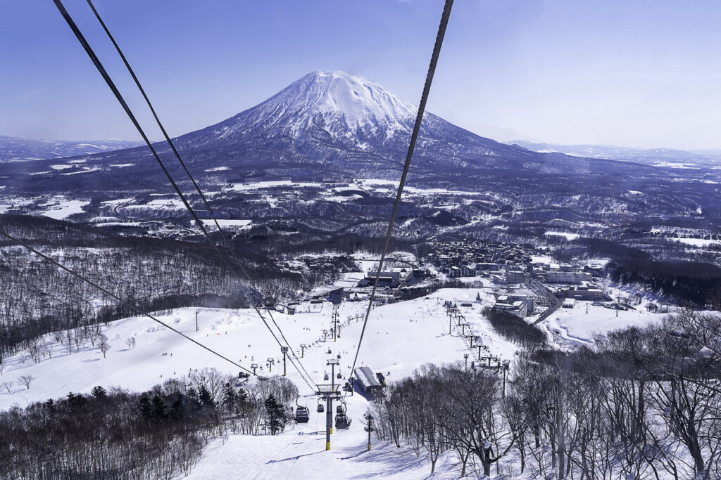 Von einer Seilbahn aus sieht man Skipisten, einen Ort im Tal und einen kegelförmigen, schneebedeckten Berg in Hokkaido.