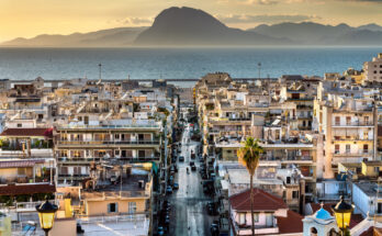 Eine gerade Straße führt durch Häuserreihen Richtung Meer mit Bergen am Horizont in Patras.