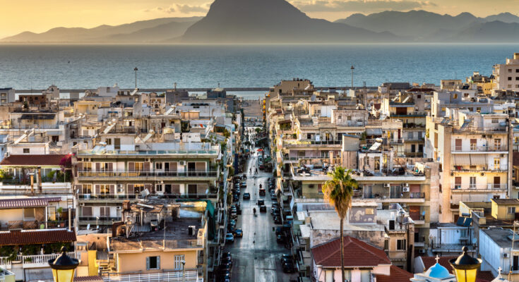 Eine gerade Straße führt durch Häuserreihen Richtung Meer mit Bergen am Horizont in Patras.