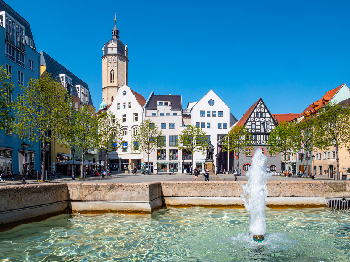 Ein Springbrunnen im Vordergrund, dahinter historische Gebäude und ein Kirchturm unter klarem Himmel.