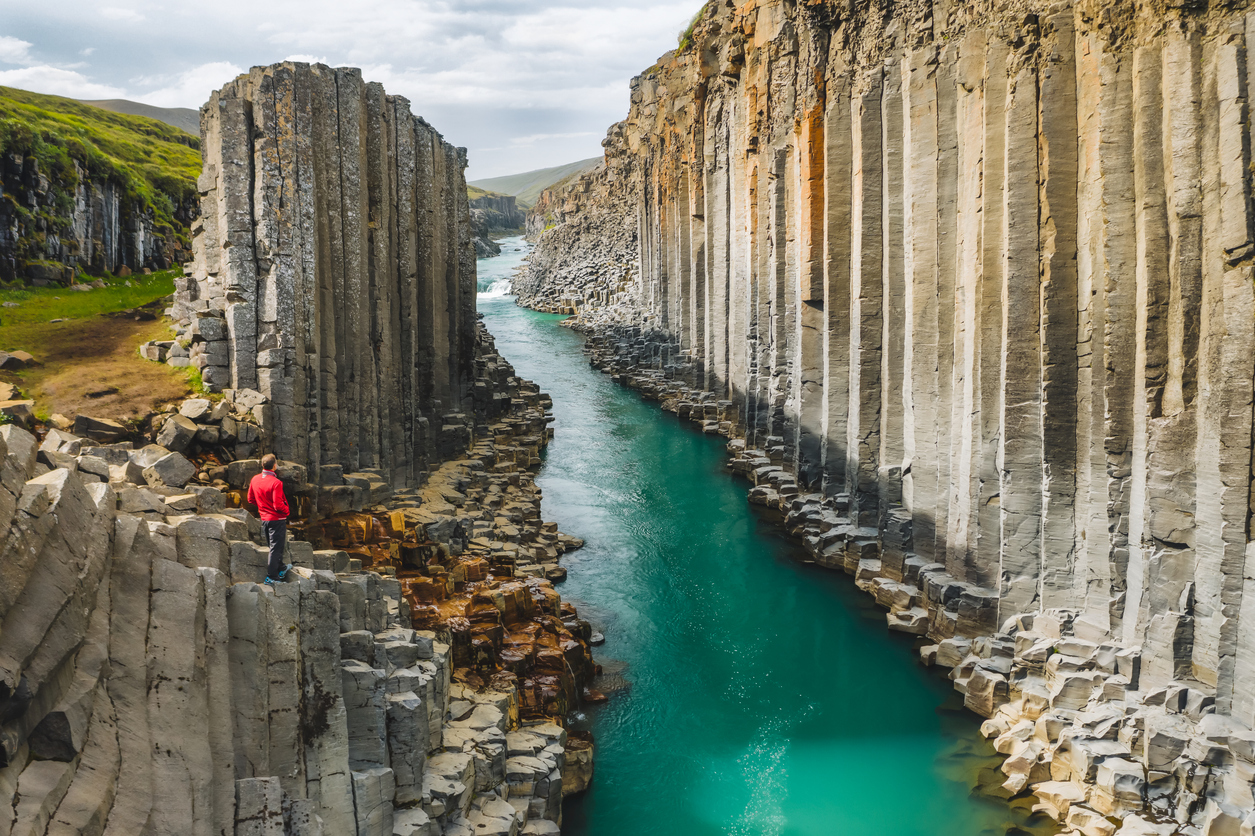 Eine Person steht in Island an einer Schlucht mit hohen Basaltsäulen, während ein türkisfarbener Fluss hindurchfließt.