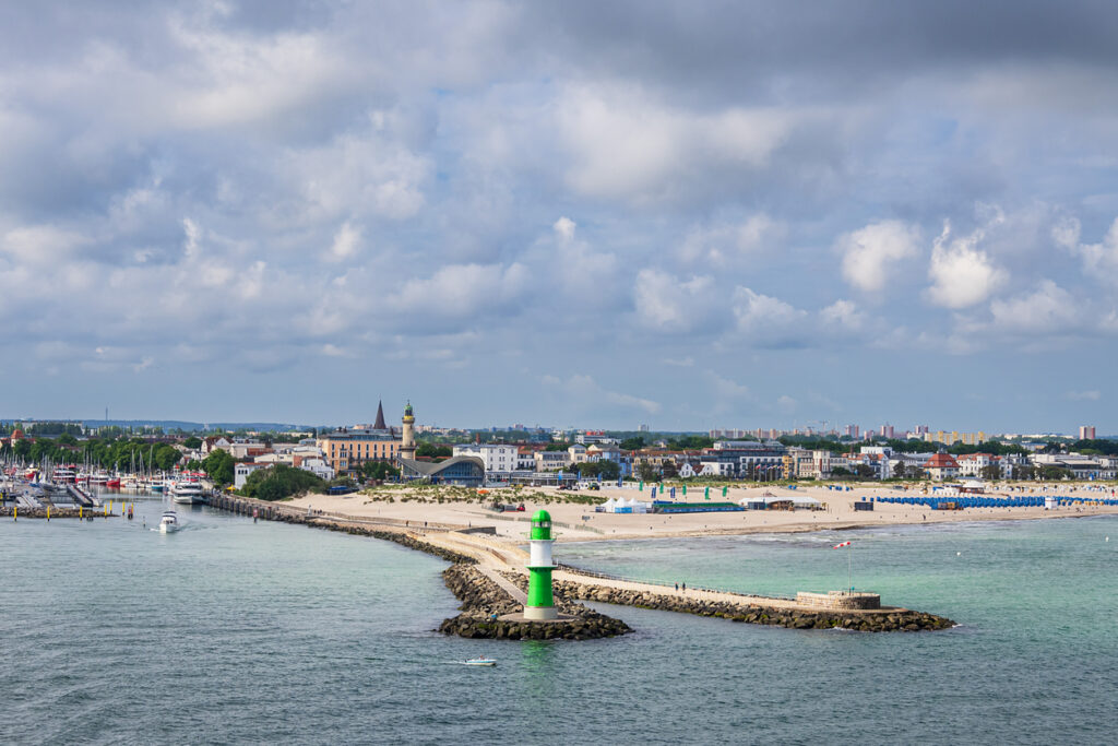 Ein grün-weißer Leuchtturm steht am Ende der Mole, dahinter liegen Strand und Stadt am Meer.