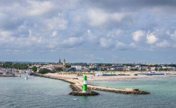 Ein grün-weißer Leuchtturm steht am Ende der Mole, dahinter liegen Strand und Stadt am Meer.
