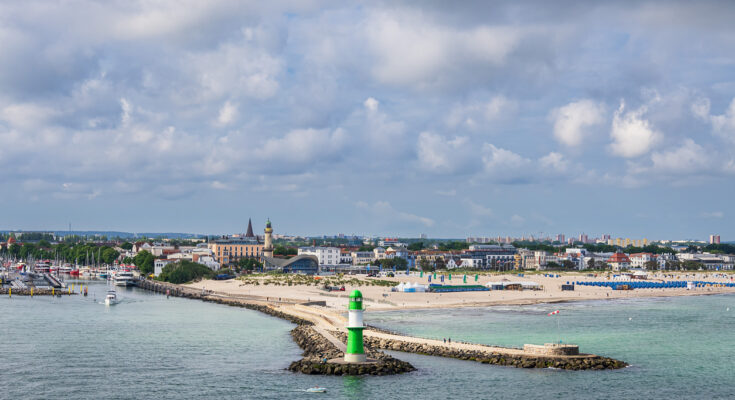 Ein grün-weißer Leuchtturm steht am Ende der Mole, dahinter liegen Strand und Stadt am Meer.