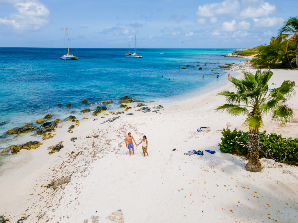 Ein Paar geht über hellen Sand, während Boote auf dem blauen Wasser vor der Küste von Aruba liegen.
