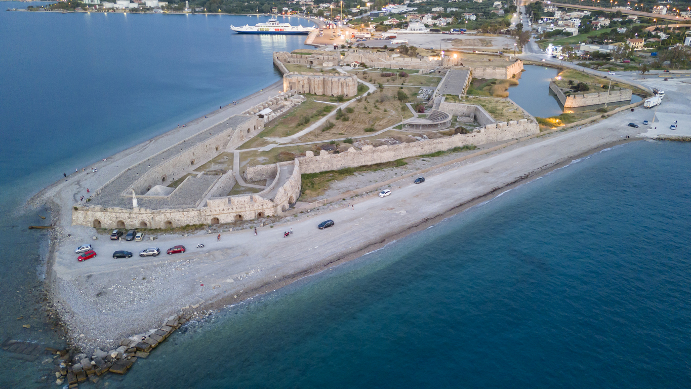 Eine lange Festungsanlage liegt an einer Landzunge am Meer, im Hintergrund sieht man den Hafen von Patras.