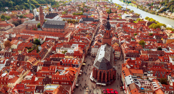 Eine Luftaufnahme zeigt dichte rote Dächer, große Kirchen und den Neckar, der sich am Stadtrand entlangzieht.