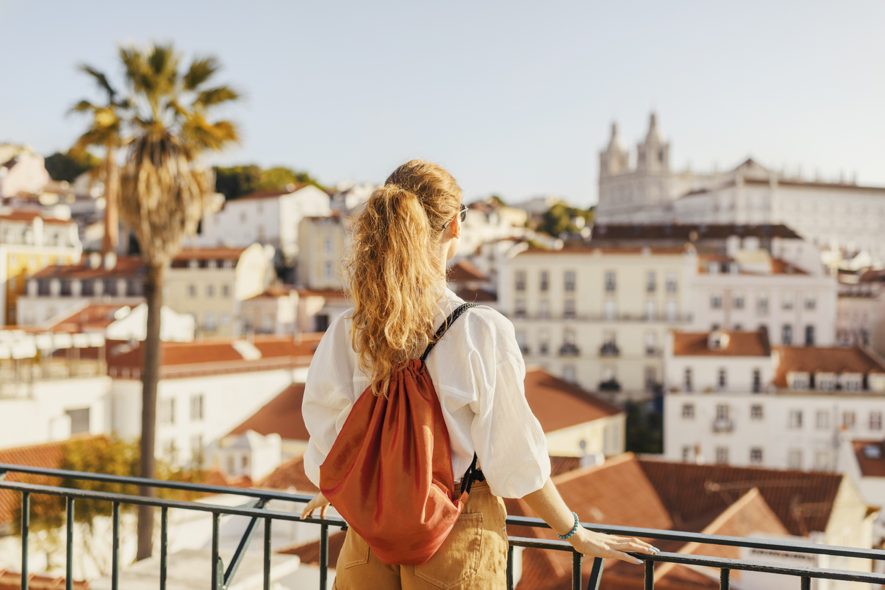 Frau mit Rucksack steht an einem Aussichtspunkt und schaut über die roten Dächer von Lissabon.