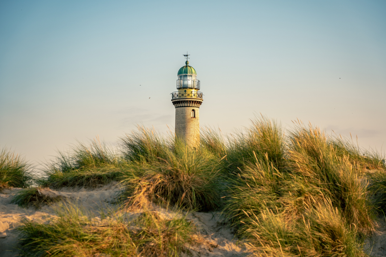 Der Leuchtturm steht hinter Dünengras, warmes Licht färbt Himmel und Sand.