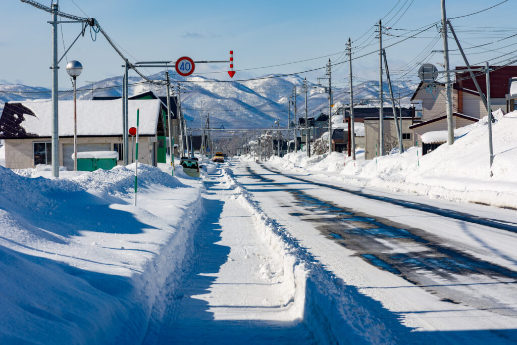 Eine geräumte Straße mit hohen Schneewällen führt durch ein Dorf in Hokkaido, dahinter liegen verschneite Berge.