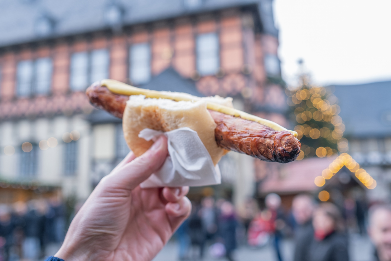 Eine Hand hält eine Bratwurst im Brötchen vor einem unscharfen Markt mit Lichtern und Fachwerk.