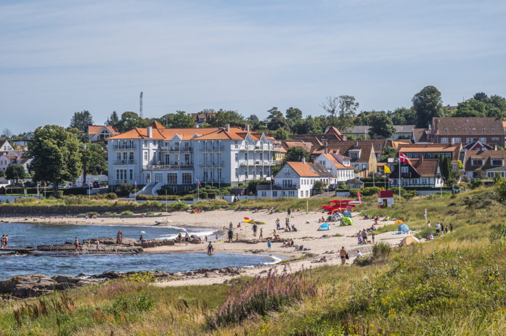 Menschen entspannen an einem Strand vor Häusern mit roten Dächern und einer Flagge auf Bornholm.
