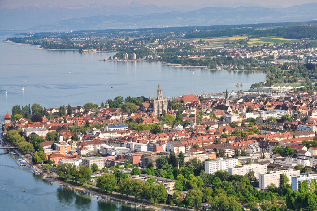 Eine Stadt mit roten Dächern liegt direkt am See, dahinter ziehen sich Felder und Hügel.