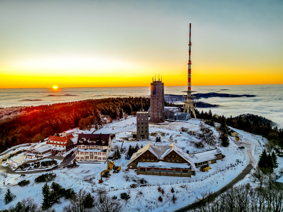 Verschneite Anlage mit Funkmast und Gebäuden bei Sonnenuntergang über einem Nebelmeer.