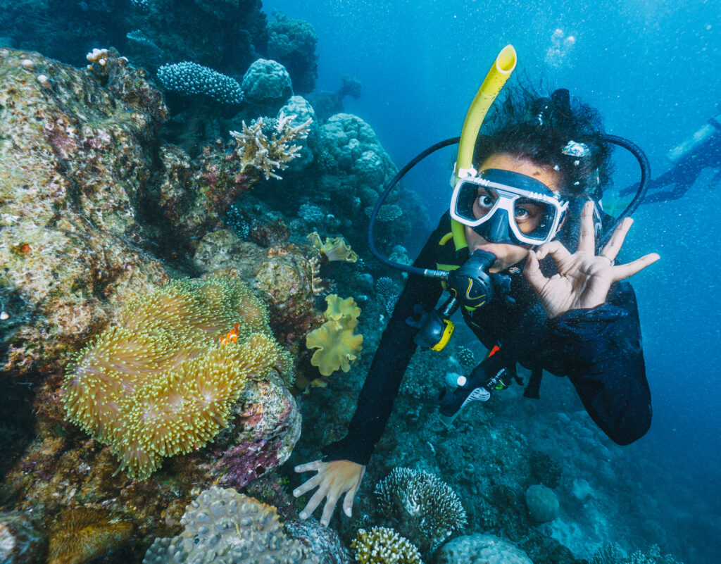 Taucherin zeigt unter Wasser ein OK-Zeichen neben bunten Korallen und einem kleinen Clownfisch.