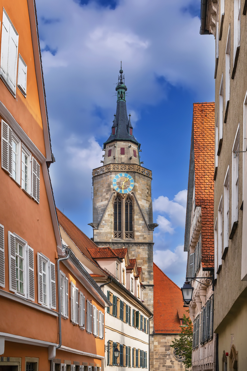 Blick durch eine schmale Gasse auf einen Kirchturm mit Uhr und rote Ziegeldächer.