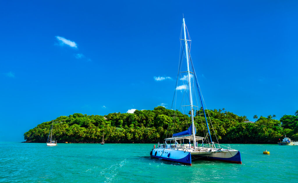 Ein Segelkatamaran fährt auf ruhigem, türkisfarbenem Wasser vor einer tropischen Insel.