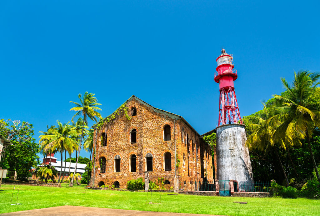 Ein roter Leuchtturm steht neben einem verwitterten Gebäude zwischen Palmen und sattem Grün.