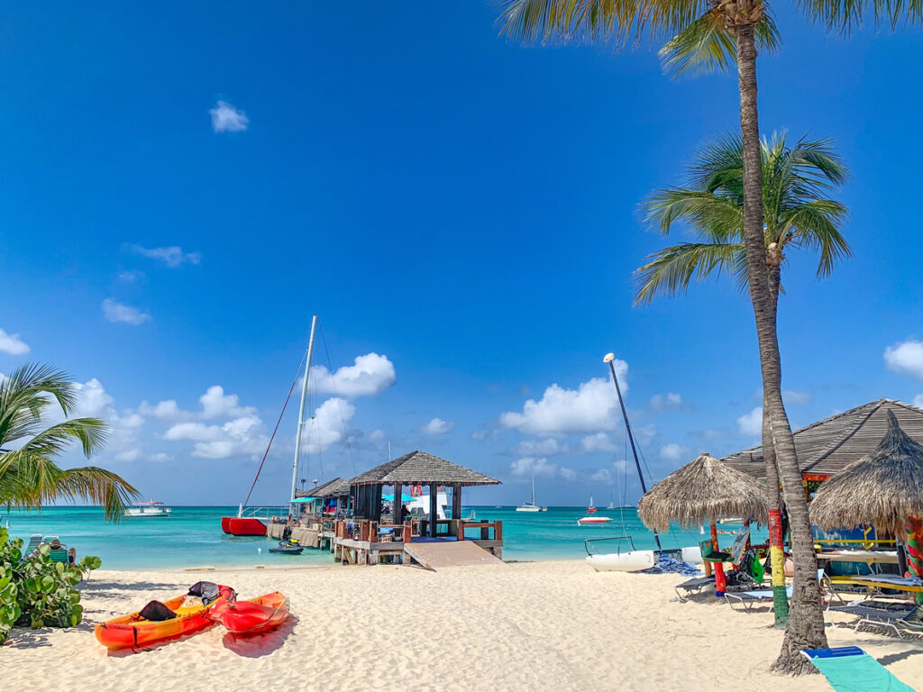 Orange Kajaks liegen im Sand, daneben ein kleiner Steg mit Hütte und Segelbooten vor Aruba.