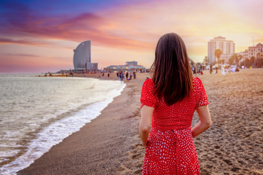 Frau im roten Kleid steht am Strand und schaut auf die Küste von Barcelona bei Sonnenuntergang.