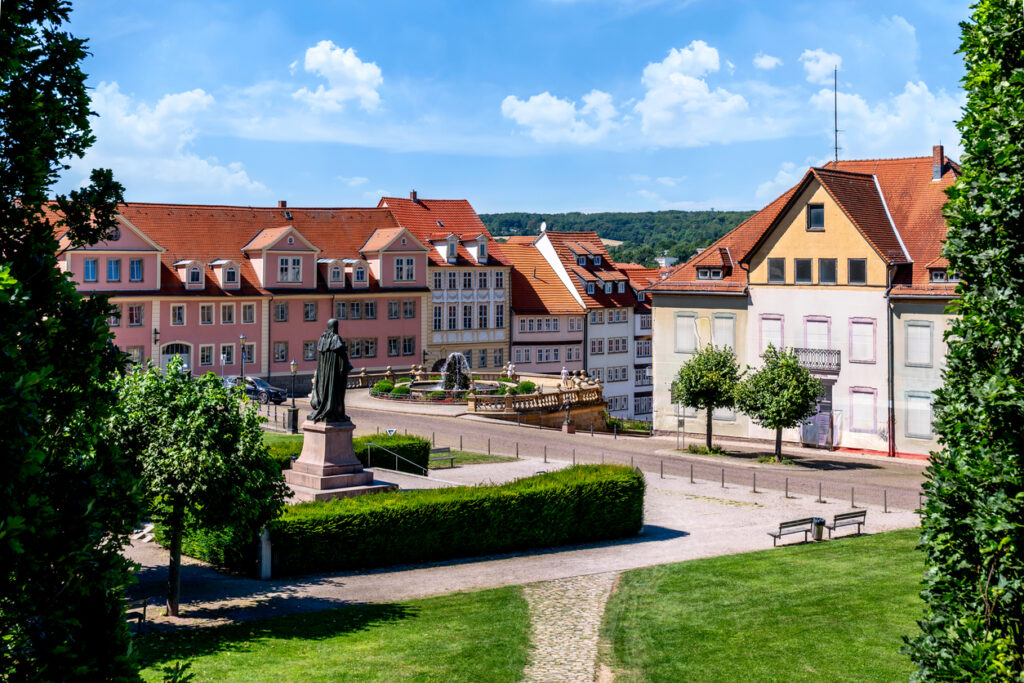 Aussicht auf einen Platz mit Statue, Brunnen und farbigen Häusern unter sommerlichem Himmel.