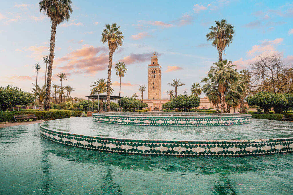 Ein runder Brunnen liegt in einem Park mit Palmen, dahinter ragt ein Minarett auf.