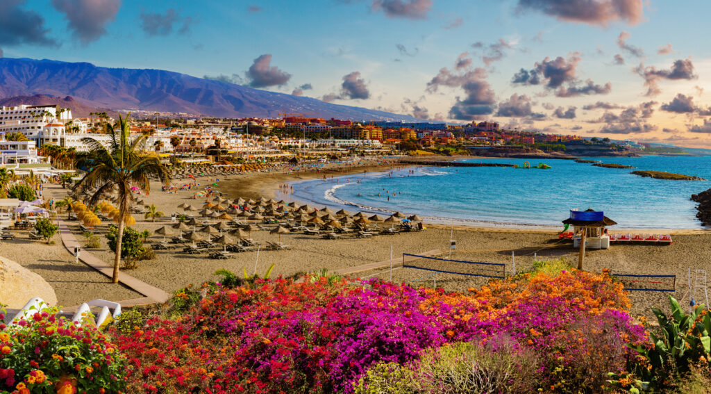 Große Strandbucht mit Sonnenschirmen, Promenade, bunten Blumen und Hotels vor Bergen