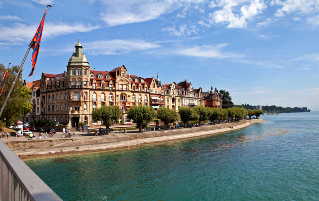 Eine Reihe historischer Gebäude steht direkt am Uferweg neben blauem Wasser.