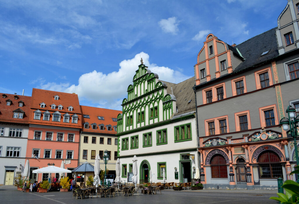 Blick auf einen Platz mit farbigen Fassaden, Außengastronomie und blauem Himmel.
