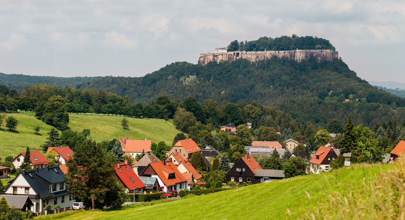 Ein Dorf mit roten Dächern liegt in einer grünen Landschaft, darüber thront die Festung Königstein auf einem bewaldeten Tafelberg.