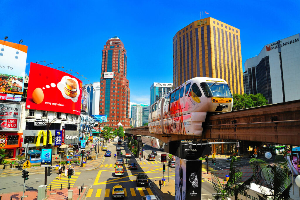 Monorail fährt über eine belebte Kreuzung mit Hochhäusern und bunten Werbetafeln in Kuala Lumpur