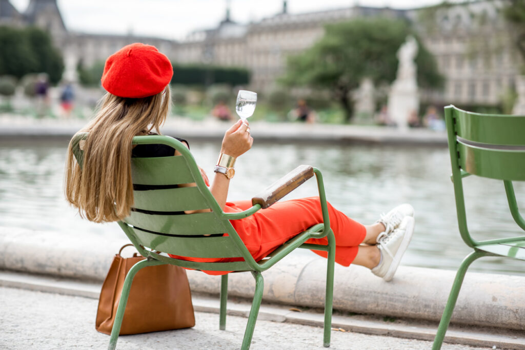 Frau mit roter Baskenmütze sitzt auf einem Stuhl im Park und hält ein Glas in Paris.