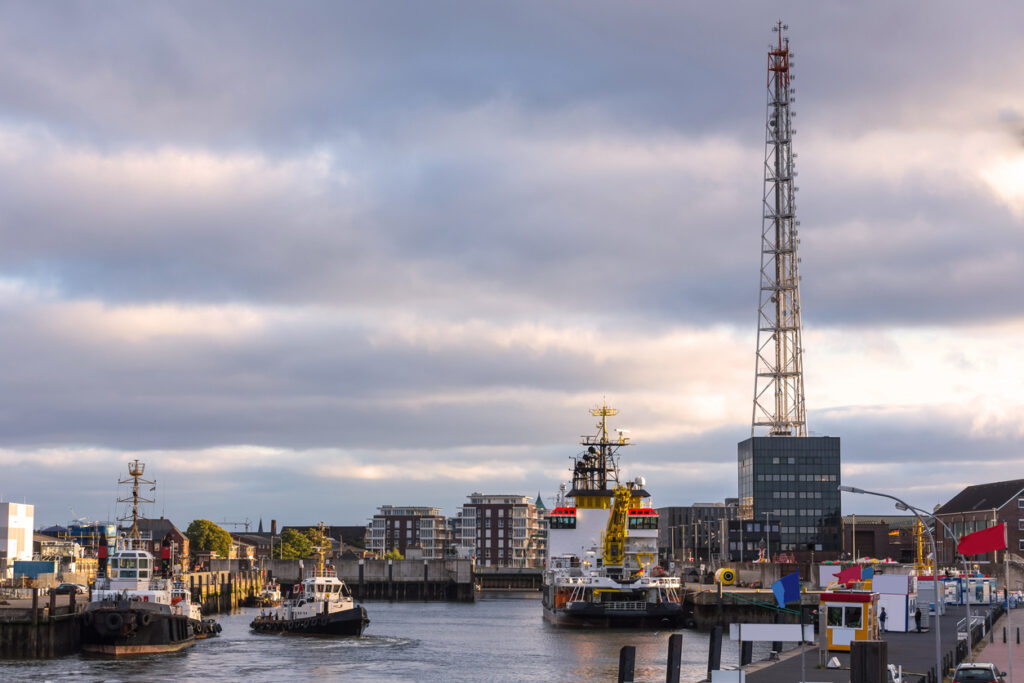 Mehrere Arbeitsboote und Schlepper liegen in einem Kanalbecken vor der Kulisse der Stadt und eines hohen Sendemasts.