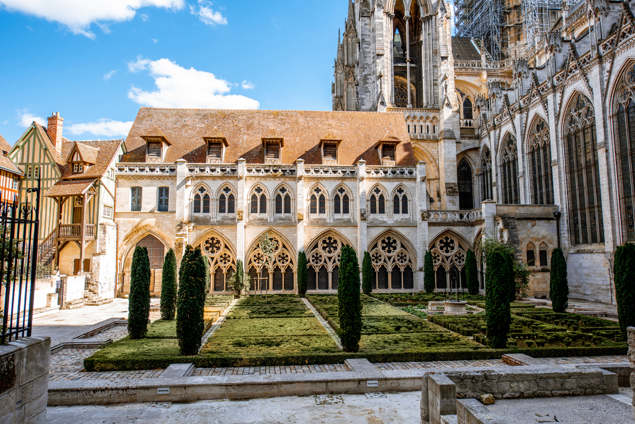 Ein sonniger Innenhof zeigt eine reich verzierte gotische Fassade mit spitzen Bögen und Maßwerkfenstern, davor akkurat geschnittene Hecken und Zypressen.
