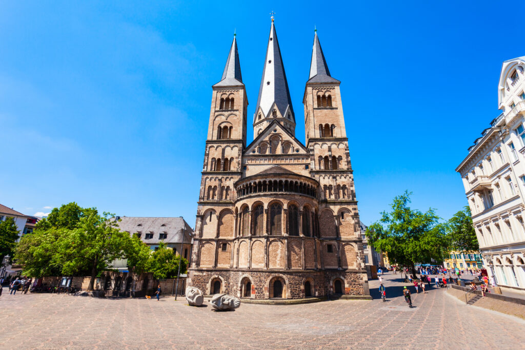 Die Rückansicht der Basilika mit ihren charakteristischen drei Türmen und der runden Apsis steht auf einem weiten, gepflasterten Platz unter blauem Himmel.