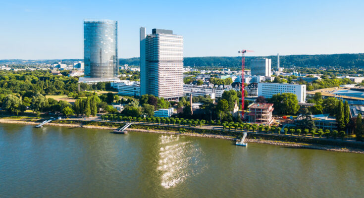 Luftaufnahme des Rheinufers mit den markanten Hochhäusern des Post Towers und des Langen Eugens unter klarem blauem Himmel.