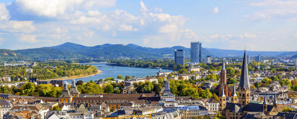 Eine weite Panoramaaufnahme zeigt im Vordergrund die Dächer der Stadt und die Türme des Münsters, dahinter den Rhein und die Silhouette der Berge am Horizont.