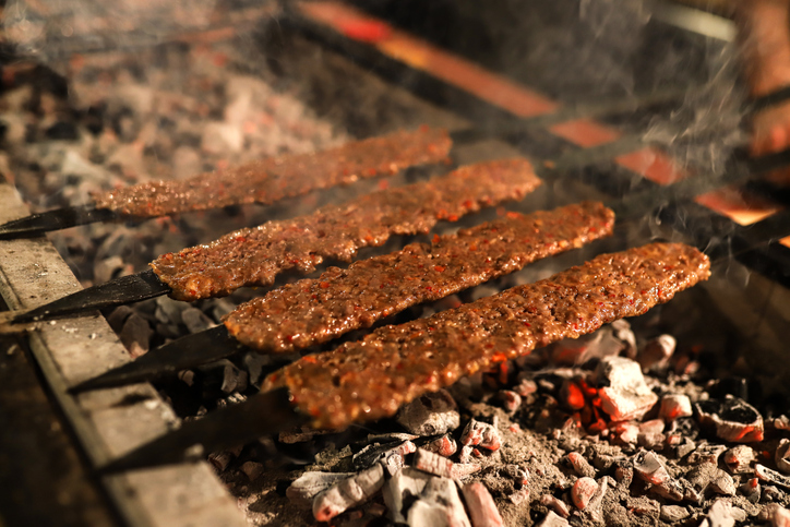 Mehrere lange Metallspieße mit Adana Kebab garen über glühender Holzkohle in einem traditionellen Grill.