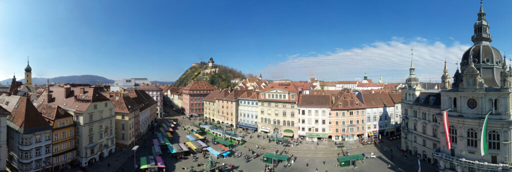 Ein 360-Grad-Panorama von Graz zeigt den belebten Hauptplatz mit Marktständen, dem Rathaus und dem Schlossberg mit Uhrturm im Hintergrund.