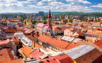 Blick über Košice mit roten Ziegeldächern, Kirchturmspitze und moderner Skyline am Horizont.