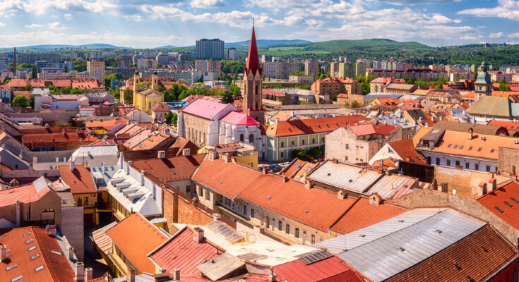 Blick über Košice mit roten Ziegeldächern, Kirchturmspitze und moderner Skyline am Horizont.