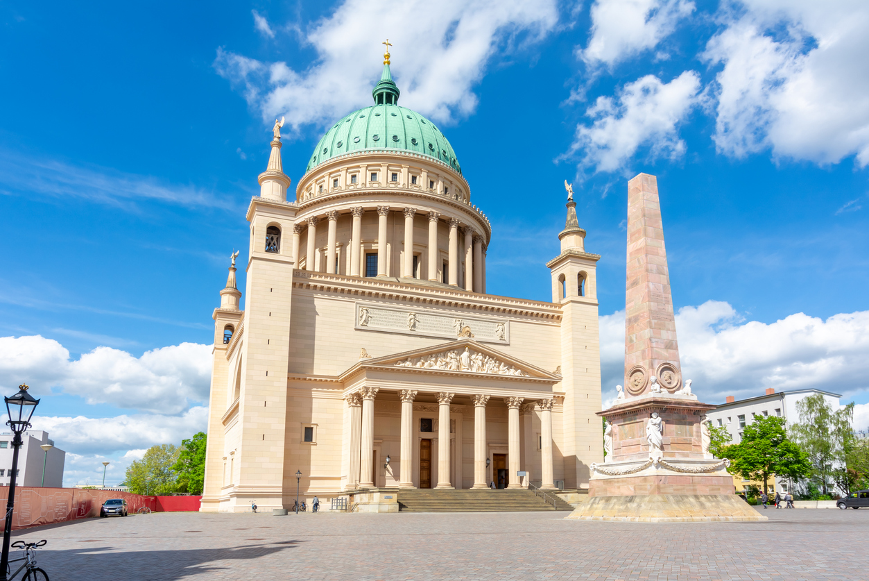 Das monumentale klassizistische Kirchengebäude mit seiner markanten grünen Kuppel steht hinter einem hohen Obelisken auf einem weiten Platz.