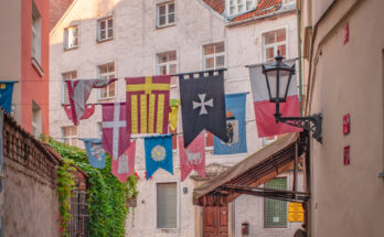 Eine schmale Kopfsteinpflastergasse in Riga ist mit bunten Wappenbannern geschmückt.