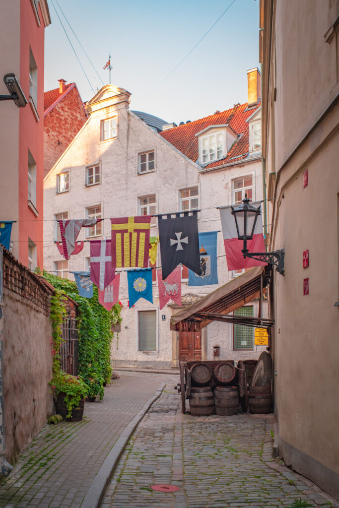 Eine schmale Kopfsteinpflastergasse in Riga ist mit bunten Wappenbannern geschmückt.