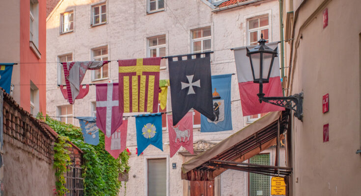 Eine schmale Kopfsteinpflastergasse in Riga ist mit bunten Wappenbannern geschmückt.