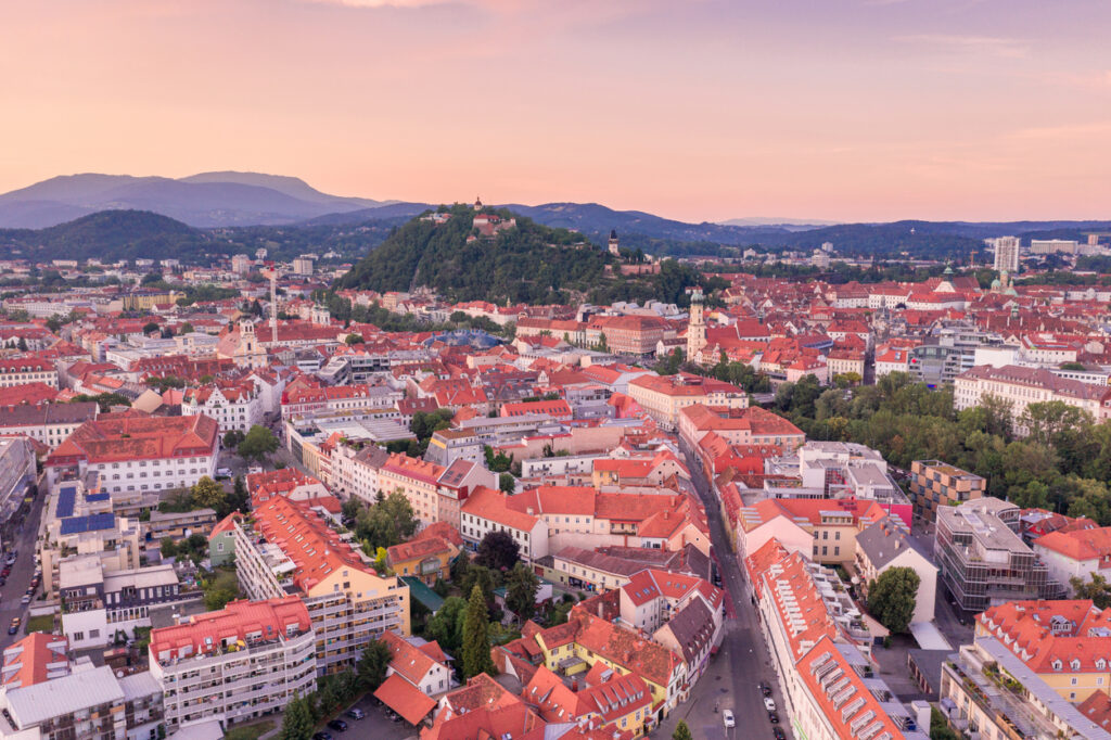 Ein weitläufiges Luftbild von Graz während der Dämmerung mit Blick auf den Schlossberg und das endlose Meer aus roten Ziegeldächern.