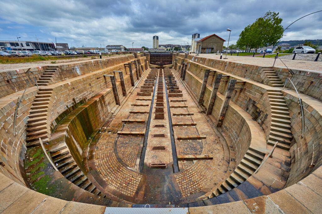 Ein tiefes, gemauertes Trockendock mit Treppenaufgängen an den Seiten unter einem dramatischen Wolkenhimmel.