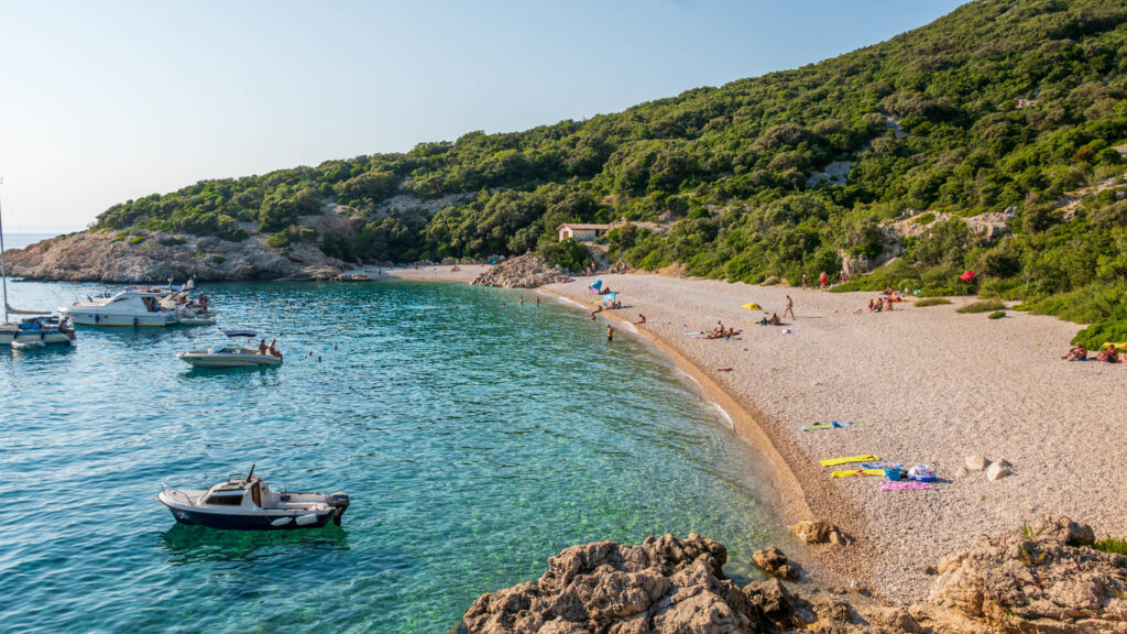 Ein heller Kieselstrand mit kristallklarem, türkisfarbenem Wasser liegt am Fuße eines steilen, grün bewachsenen Hangs; kleine Motorboote ankern in der Bucht.
