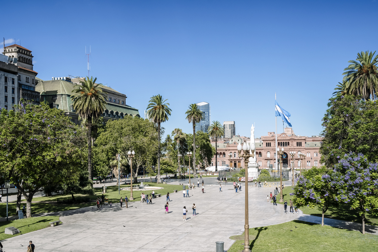 Ein weitläufiger Platz in Buenos Aires mit Spaziergängern, Palmen und dem rosa Regierungsgebäude im Hintergrund unter blauem Himmel.